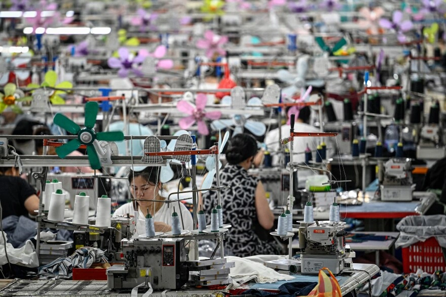 Workers operate sewing machines in a large, crowded textile factory filled with colorful fans and fabric spools.