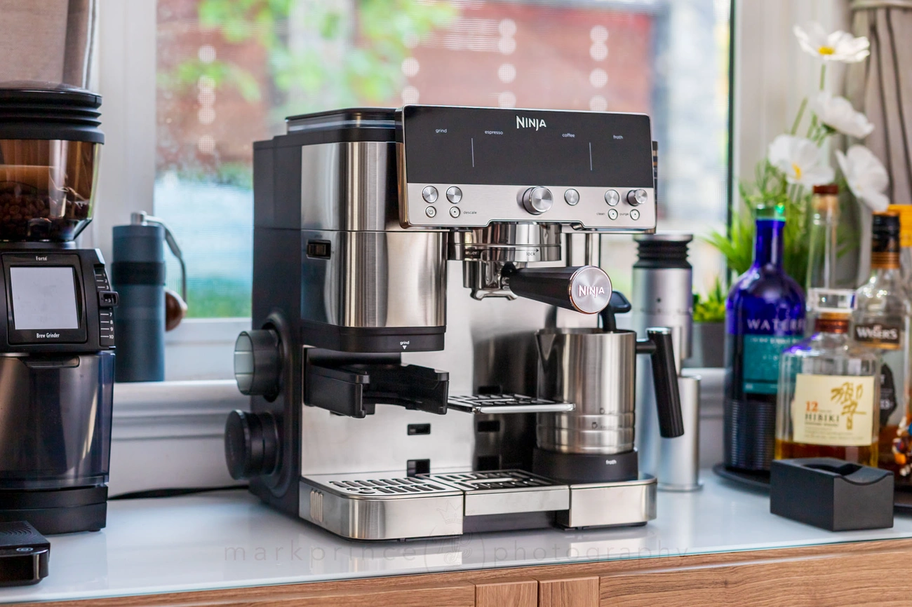 Stainless steel Ninja espresso machine on kitchen counter with grinder and assorted bottles in background.