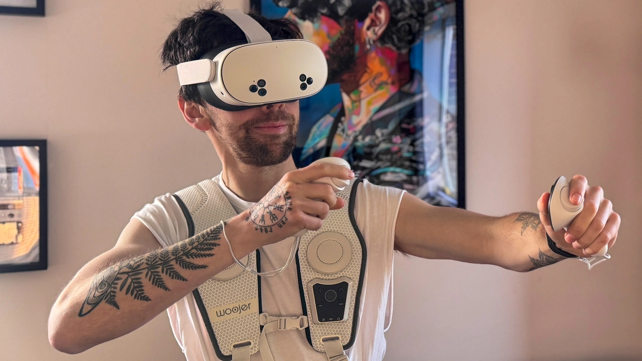 Person wearing a VR headset and a Woojer haptic vest, holding VR controllers while immersed in virtual reality.