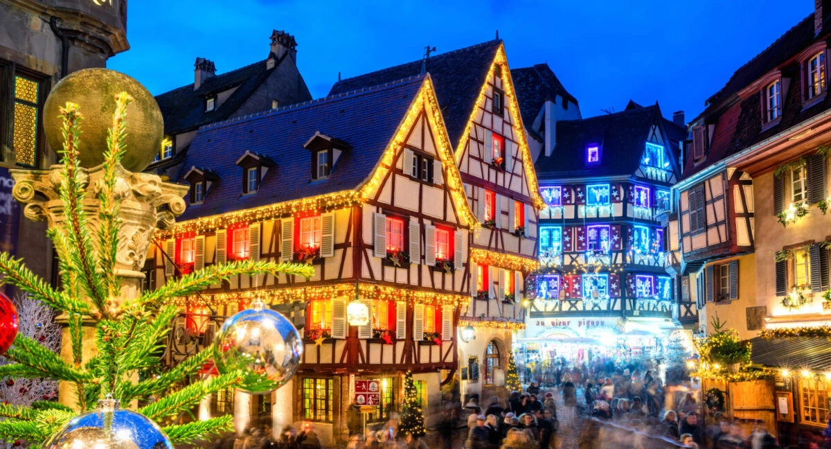 Festively lit half-timbered houses and a bustling Christmas market crowd in Colmar, France at dusk.