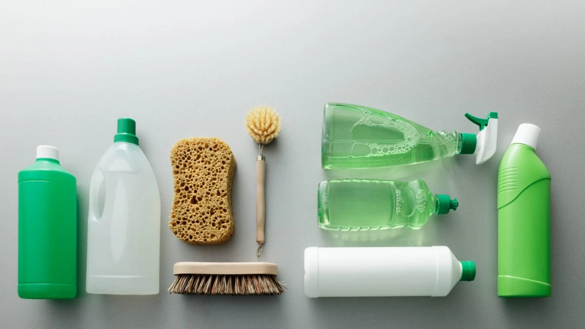 Various green and natural cleaning bottles, brushes, and sponges arranged on a gray surface promoting eco-friendly cleaning.