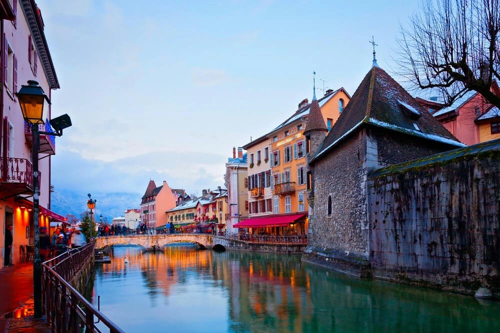 Scenic view of Annecy's old town canal at dusk with colorful buildings and a stone bridge reflecting in the water.