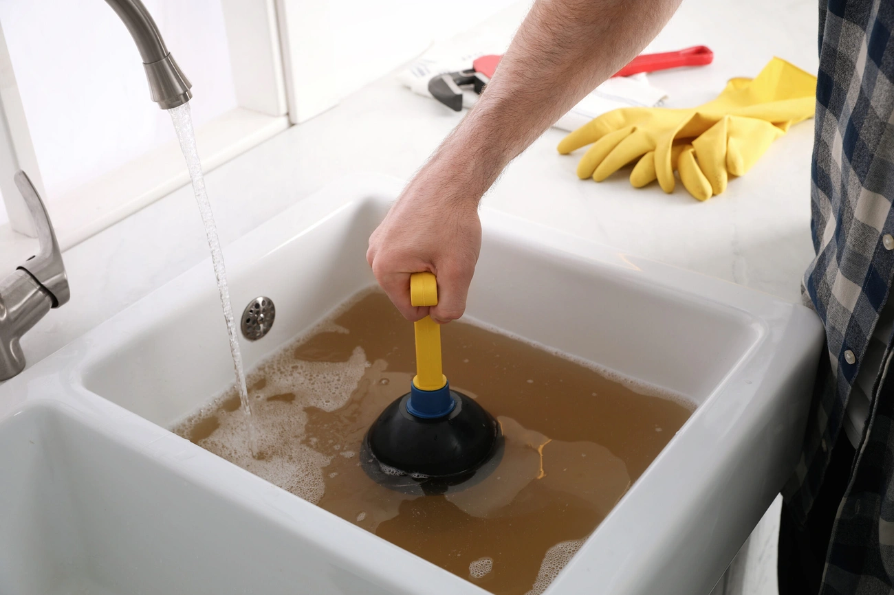Person using a plunger to unclog a sink filled with dirty water and running faucet water.