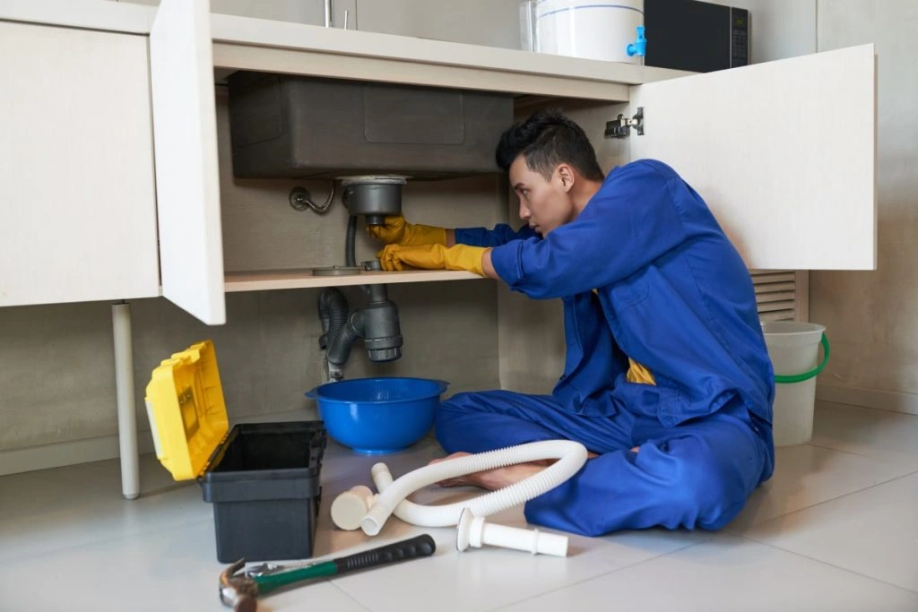 Plumber in blue coveralls fixing a kitchen sink drain with tools and pipes on the floor nearby.