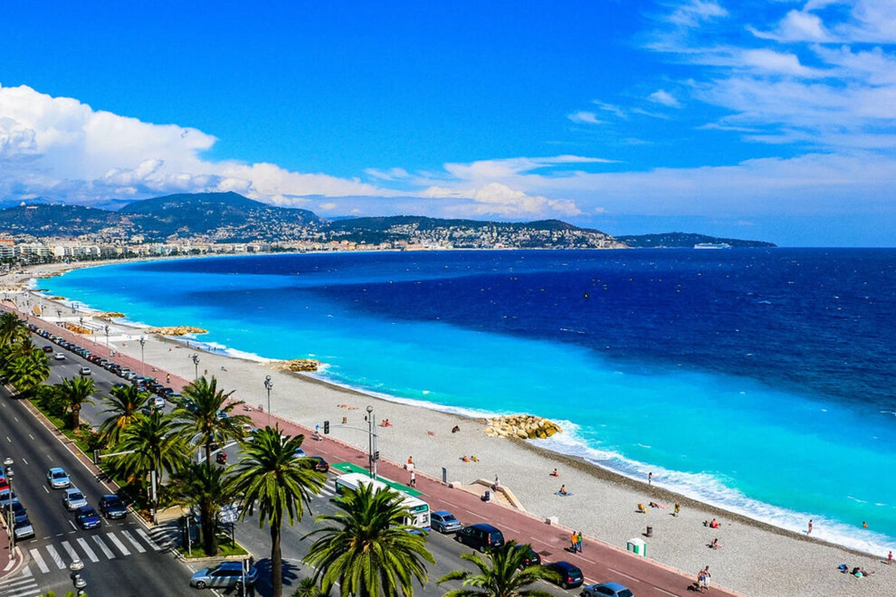 Aerial view of Nice, France coastline with palm-lined road, pebble beach, and blue Mediterranean Sea under a sunny sky.