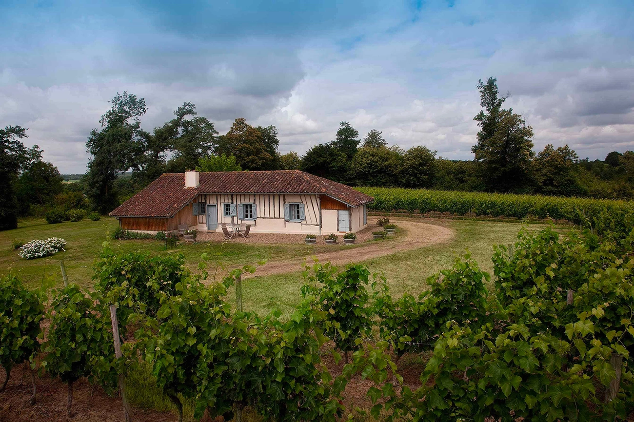 Traditional countryside house surrounded by green vineyards and trees under a cloudy sky in Landes, France.