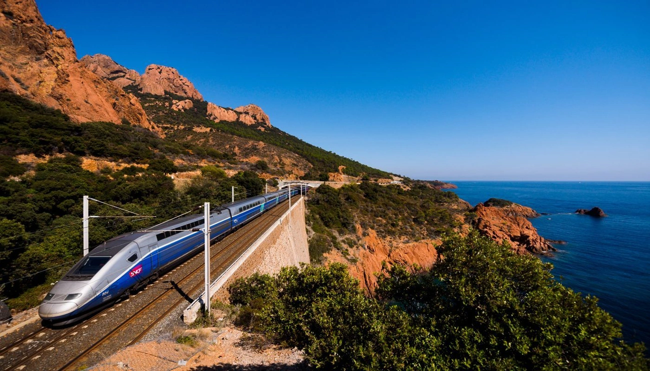 High-speed train traveling along a coastal railway with rocky cliffs and blue sea under a clear sky in France.