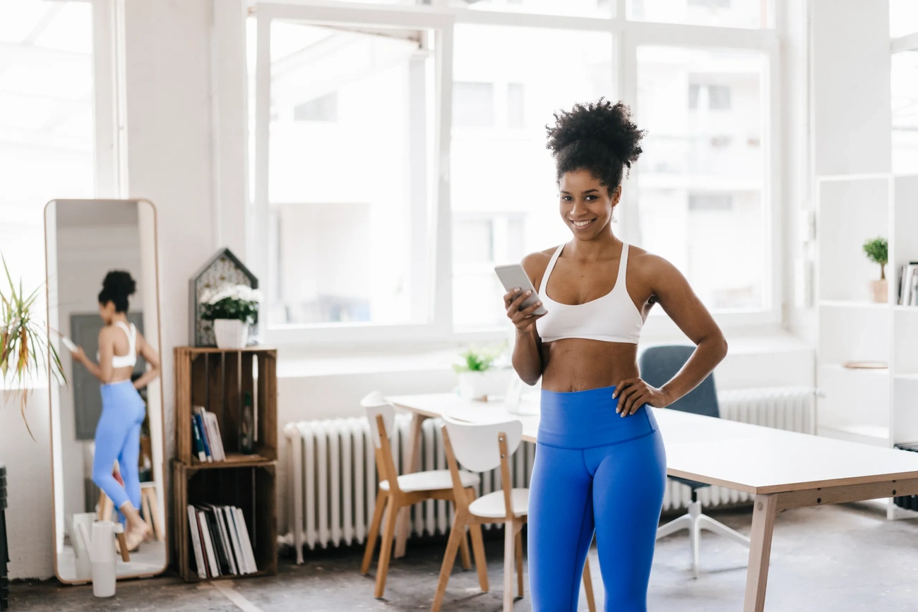 Woman in blue leggings and white sports bra holding a phone in a bright room with large windows and furniture.