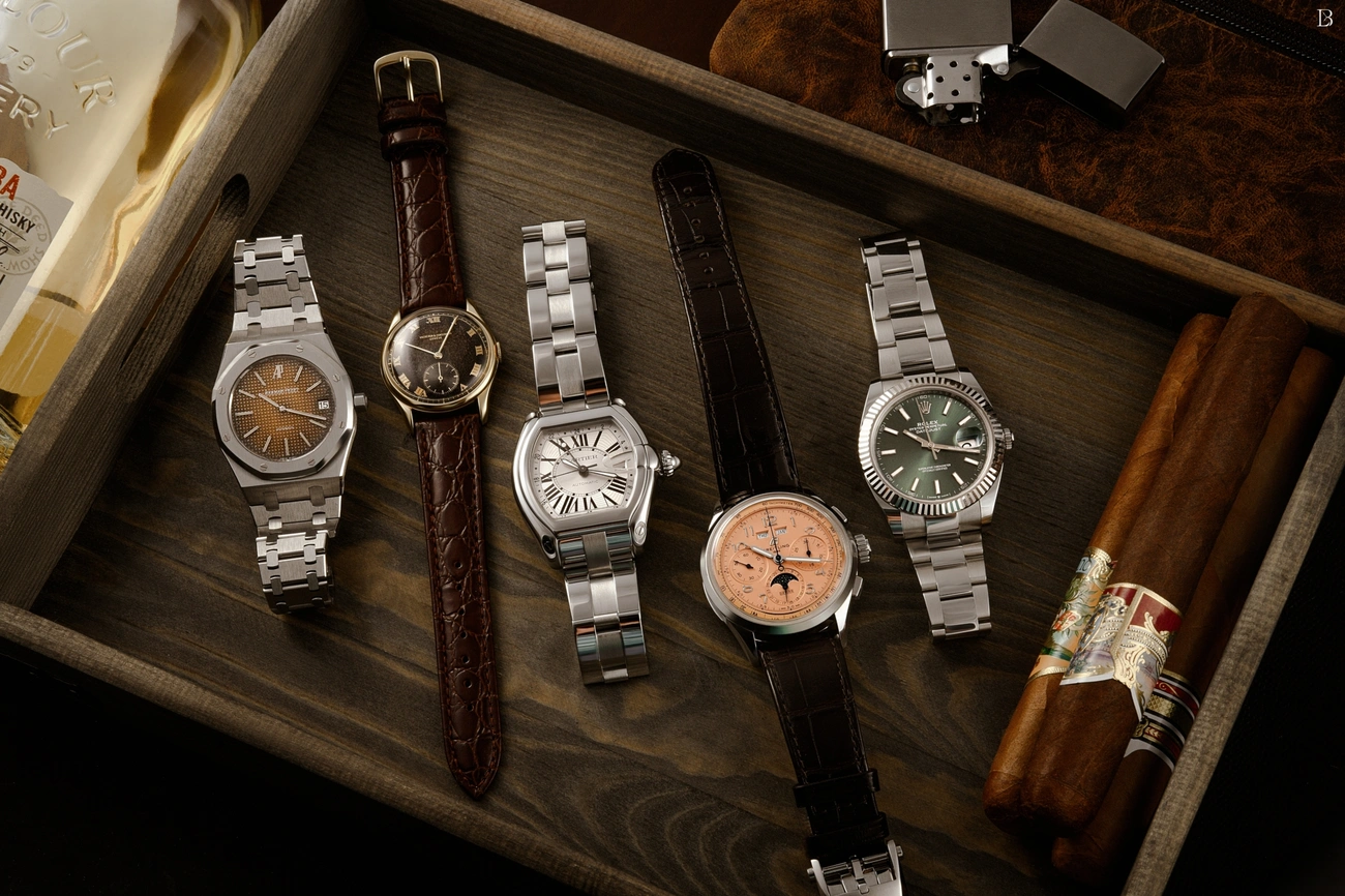 Five luxury wristwatches with metal and leather bands displayed in a wooden tray alongside cigars and a lighter.