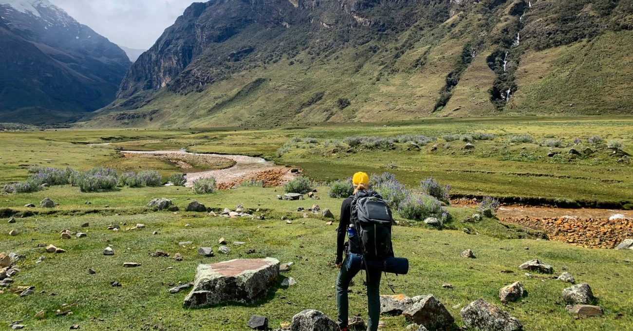 A hiker with a backpack walks through a green valley with mountains and a winding creek in the background.